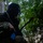 Multi-Domain Reconnaissance Course| Reconnaissance and Surveillance Exercise A soldier in camouflage uniform and insect/mesh head net is positioned in a wooded area, holding a rifle with a blue training magazine. The image is heavily shadowed, with the subject largely in silhouette against a green forest background. The blue magazine indicates this is likely a training exercise rather than a live combat situation.