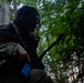 A soldier in camouflage uniform and insect/mesh head net is positioned in a wooded area, holding a rifle with a blue training magazine. The image is heavily shadowed, with the subject largely in silhouette against a green forest background. The blue magazine indicates this is likely a training exercise rather than a live combat situation.