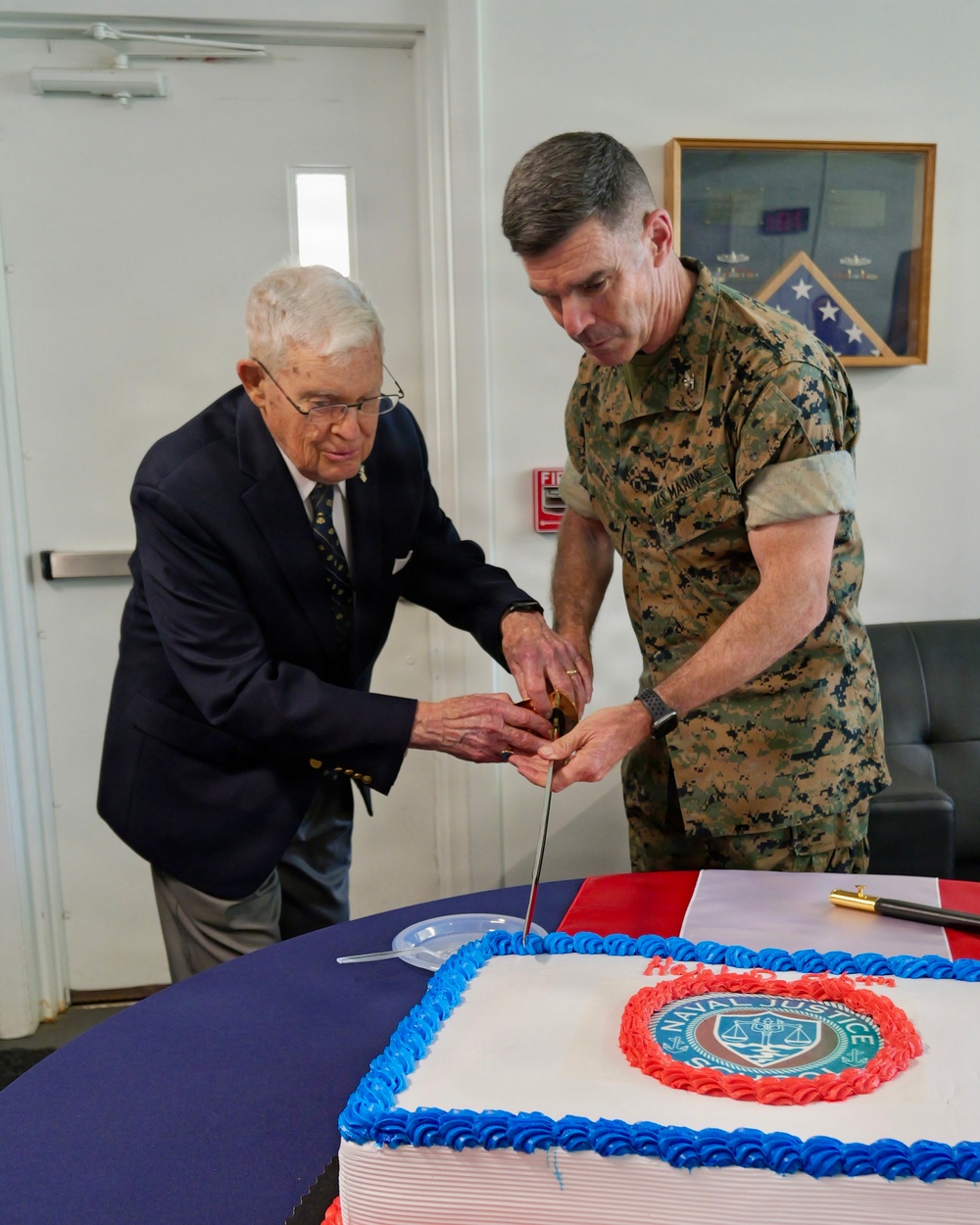Two individuals are cutting a large ceremonial cake together at what appears to be a Naval Justice School anniversary or celebration event. One person is an elderly civilian in a dark suit, and the other is a U.S. Marine Corps officer in MARPAT camouflage uniform. They are using a ceremonial sword or saber to cut the cake, which bears the Naval Justice School seal.