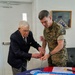 Two individuals are cutting a large ceremonial cake together at what appears to be a Naval Justice School anniversary or celebration event. One person is an elderly civilian in a dark suit, and the other is a U.S. Marine Corps officer in MARPAT camouflage uniform. They are using a ceremonial sword or saber to cut the cake, which bears the Naval Justice School seal.