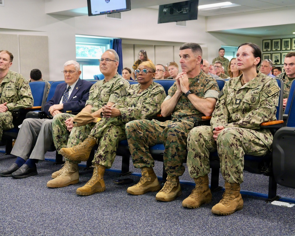 A group of U.S. Navy personnel in digital camouflage uniforms (NWU Type III) are seated in an auditorium or briefing room alongside one civilian in a suit. They appear to be attending a formal briefing, presentation, or ceremony, all facing the same direction. The setting appears to be a military installation conference or lecture facility.