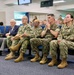 A group of U.S. Navy personnel in digital camouflage uniforms (NWU Type III) are seated in an auditorium or briefing room alongside one civilian in a suit. They appear to be attending a formal briefing, presentation, or ceremony, all facing the same direction. The setting appears to be a military installation conference or lecture facility.