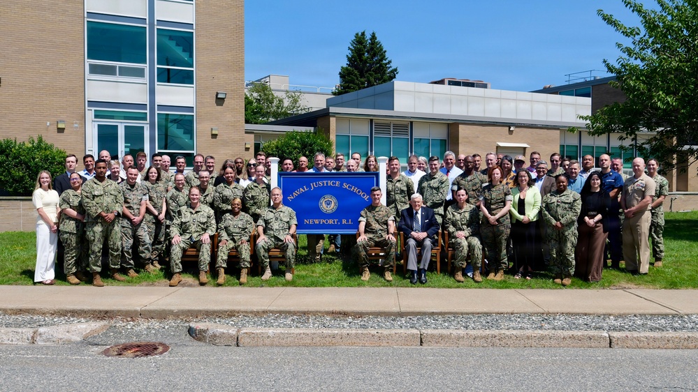 A large group photo taken in front of the Naval Justice School in Newport, Rhode Island. The group consists of a mix of military personnel in camouflage uniforms and civilians, posed in multiple rows on a grassy area. A blue sign reading 'Naval Justice School, Newport, R.I.' is prominently displayed in the center of the group.