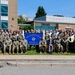 A large group photo taken in front of the Naval Justice School in Newport, Rhode Island. The group consists of a mix of military personnel in camouflage uniforms and civilians, posed in multiple rows on a grassy area. A blue sign reading 'Naval Justice School, Newport, R.I.' is prominently displayed in the center of the group.