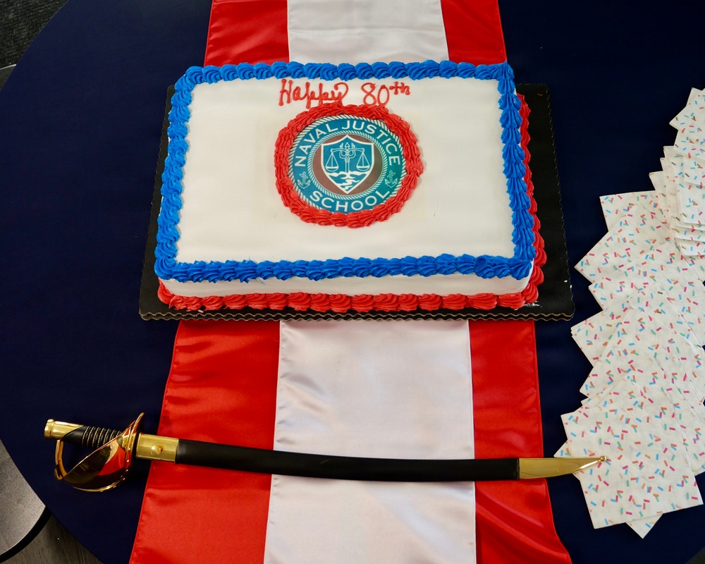 A celebratory cake for the Naval Justice School's 80th anniversary is displayed on a red and white striped table runner on a navy blue table. The rectangular white cake features blue and red frosting borders, the Naval Justice School seal in the center, and 'Happy 80th' written in red script. A ceremonial naval officer's sword with a gold and black handle is placed in front of the cake, alongside a stack of festive napkins.