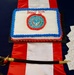 A celebratory cake for the Naval Justice School's 80th anniversary is displayed on a red and white striped table runner on a navy blue table. The rectangular white cake features blue and red frosting borders, the Naval Justice School seal in the center, and 'Happy 80th' written in red script. A ceremonial naval officer's sword with a gold and black handle is placed in front of the cake, alongside a stack of festive napkins.