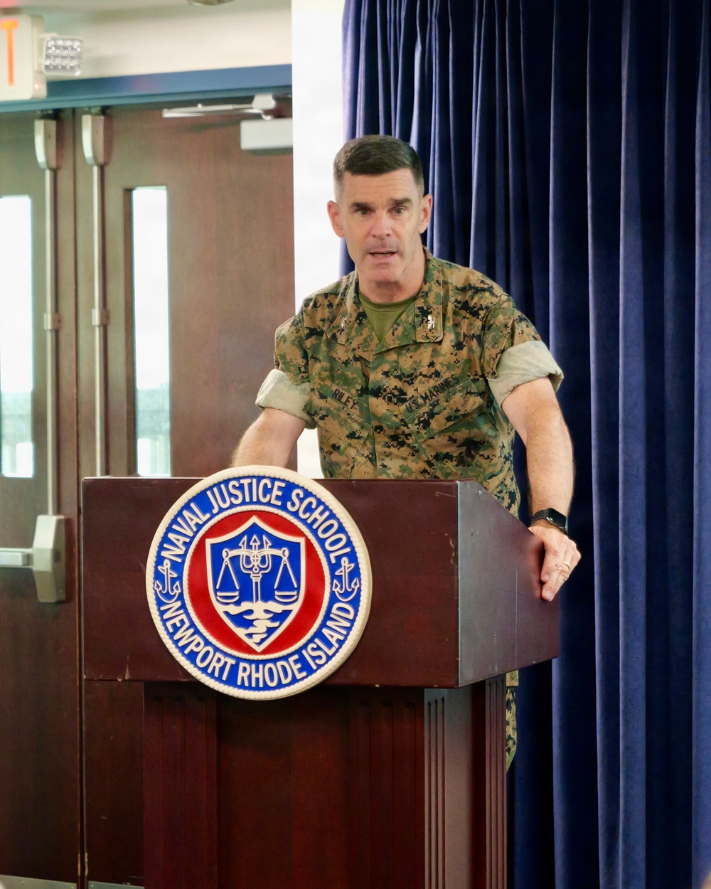 A U.S. Marine Corps officer in MARPAT camouflage uniform is speaking at a podium at the Naval Justice School in Newport, Rhode Island. The officer's name tape reads 'RILEY' and the uniform includes 'U.S. MARINES' tape. The setting appears to be a formal presentation or lecture in an auditorium-style room.