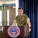 A U.S. Marine Corps officer in MARPAT camouflage uniform is speaking at a podium at the Naval Justice School in Newport, Rhode Island. The officer's name tape reads 'RILEY' and the uniform includes 'U.S. MARINES' tape. The setting appears to be a formal presentation or lecture in an auditorium-style room.