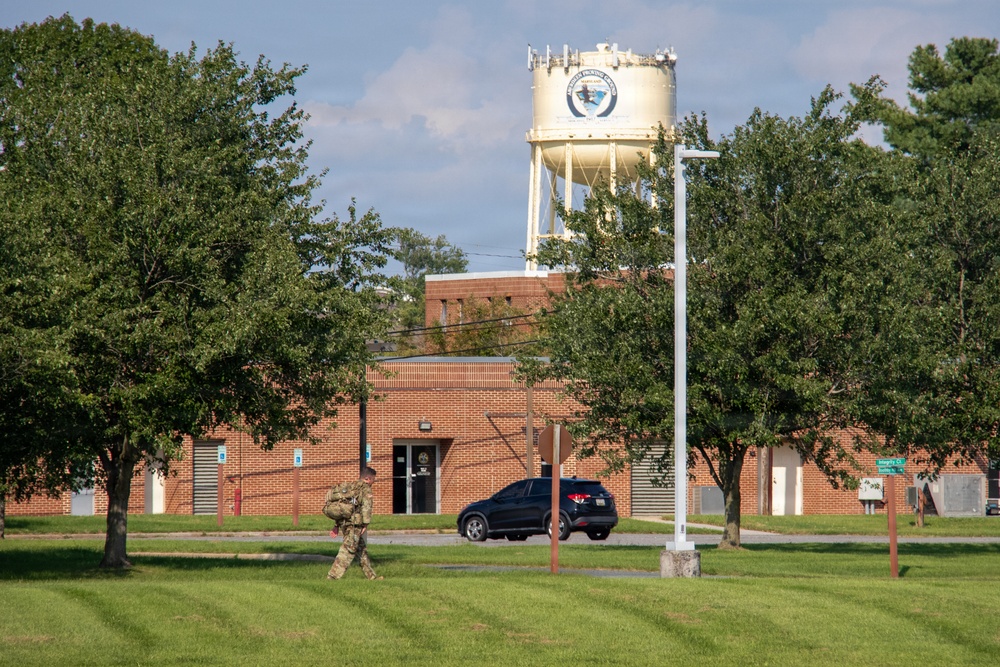 The image shows a military installation or government facility featuring a brick building and a prominent water tower displaying an official seal or emblem. A soldier in camouflage uniform carrying gear is walking across a well-maintained grassy area in the foreground. A dark-colored SUV is parked near the building entrance.