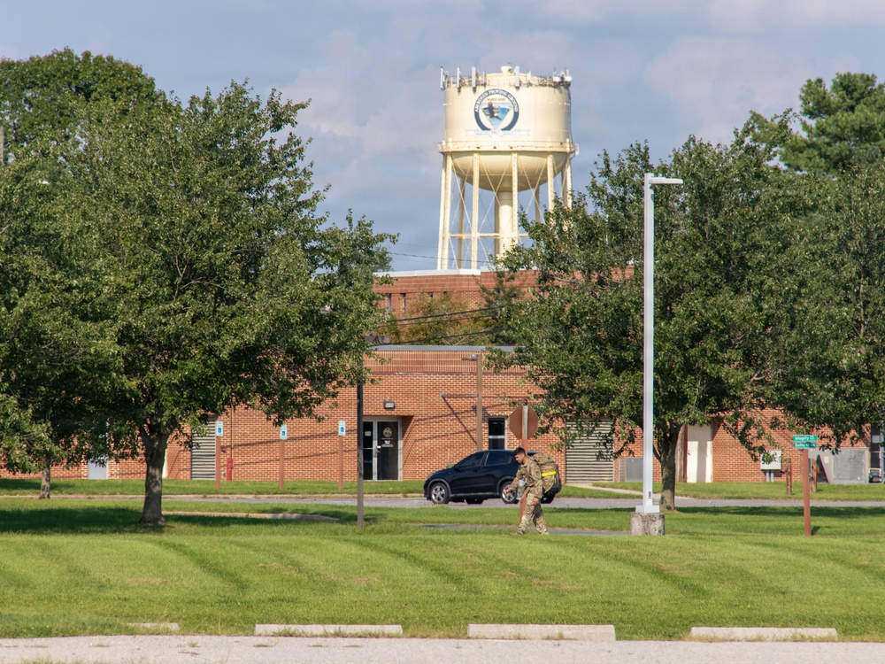 The image shows a military or government installation featuring a brick building with a large water tower bearing an official seal or emblem in the background. A person in military camouflage uniform is walking across a well-maintained grassy area near a parked dark SUV. The scene appears to be taken during daytime with partly cloudy skies.