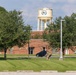 The image shows a military or government installation featuring a brick building with a large water tower bearing an official seal or emblem in the background. A person in military camouflage uniform is walking across a well-maintained grassy area near a parked dark SUV. The scene appears to be taken during daytime with partly cloudy skies.