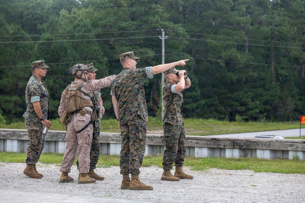 A group of four military personnel in U.S. Marine Corps MARPAT camouflage uniforms are gathered outdoors near a guardrail on what appears to be a military installation or training area. One service member is pointing in a specific direction while another uses binoculars to observe the indicated area. The setting includes a paved road, gravel surface, power lines, and a tree line in the background.
