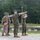 BLT 1/8 conduct organic munitions range A group of four military personnel in U.S. Marine Corps MARPAT camouflage uniforms are gathered outdoors near a guardrail on what appears to be a military installation or training area. One service member is pointing in a specific direction while another uses binoculars to observe the indicated area. The setting includes a paved road, gravel surface, power lines, and a tree line in the background.