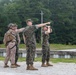 A group of four military personnel in U.S. Marine Corps MARPAT camouflage uniforms are gathered outdoors near a guardrail on what appears to be a military installation or training area. One service member is pointing in a specific direction while another uses binoculars to observe the indicated area. The setting includes a paved road, gravel surface, power lines, and a tree line in the background.