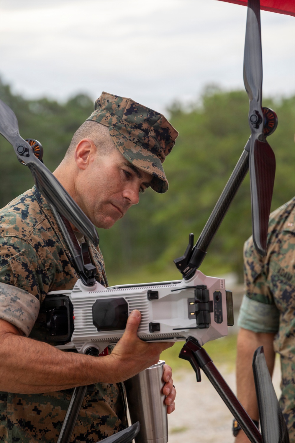 A U.S. Marine in MARPAT camouflage uniform is inspecting or handling a large multi-rotor drone, examining its underside closely. The setting appears to be an outdoor training or demonstration area with a sandy ground and tree line in the background. A second person in similar military uniform is partially visible on the right side of the frame.