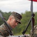 A U.S. Marine in MARPAT camouflage uniform is inspecting or handling a large multi-rotor drone, examining its underside closely. The setting appears to be an outdoor training or demonstration area with a sandy ground and tree line in the background. A second person in similar military uniform is partially visible on the right side of the frame.