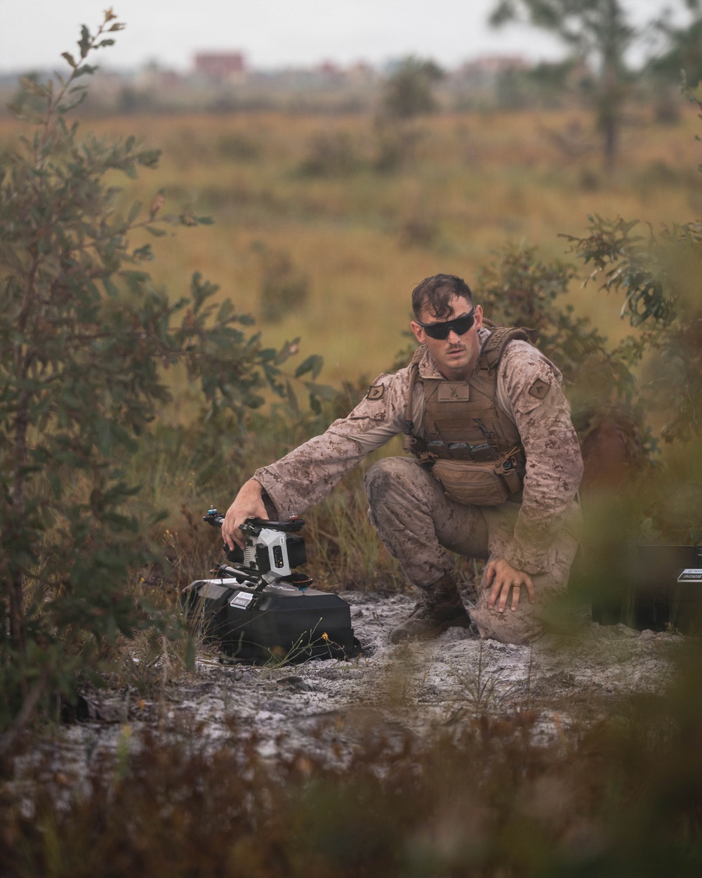 A military service member in desert MARPAT camouflage uniform is crouched in an open field environment, handling what appears to be a small unmanned ground vehicle (UGV) or robotic system placed on a black carrying case. The individual is wearing tactical gear including a plate carrier vest, sunglasses, and appears to be wet or sweating. The setting suggests a field training exercise in a scrubland or open terrain environment.