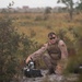 A military service member in desert MARPAT camouflage uniform is crouched in an open field environment, handling what appears to be a small unmanned ground vehicle (UGV) or robotic system placed on a black carrying case. The individual is wearing tactical gear including a plate carrier vest, sunglasses, and appears to be wet or sweating. The setting suggests a field training exercise in a scrubland or open terrain environment.