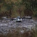 A quadcopter drone is positioned on muddy, wet ground surrounded by sparse grass and shrubs. The drone appears to be either landing or taking off, as its propellers appear to be spinning (motion blur visible). The setting is an outdoor, semi-wild terrain with overcast lighting conditions.