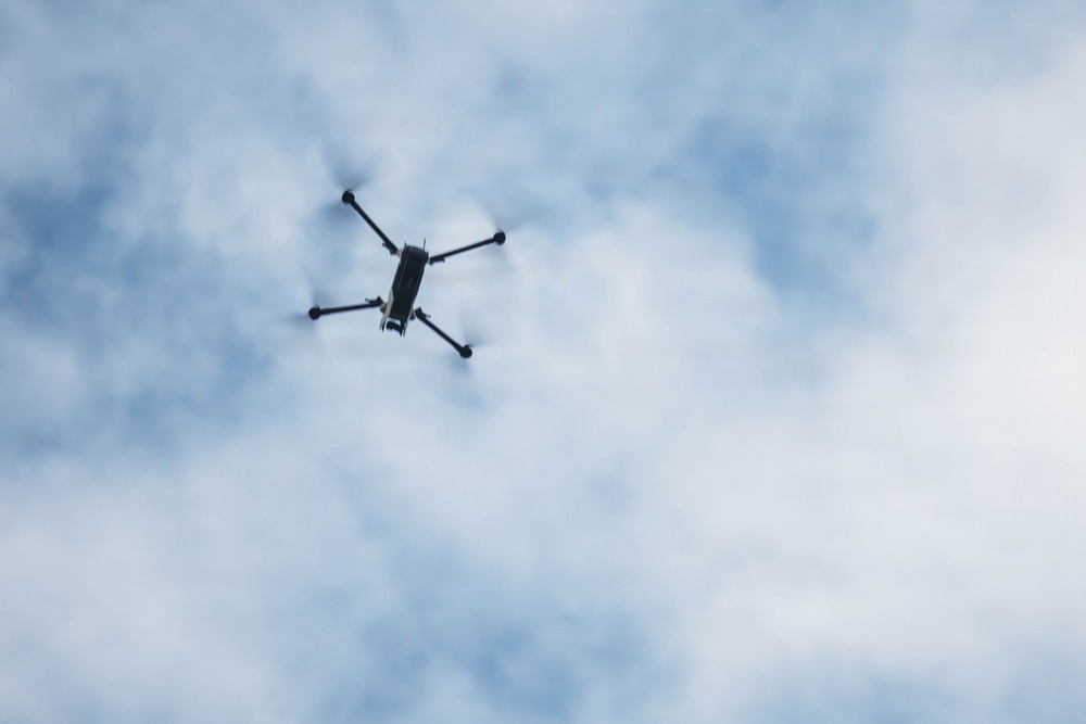 A quadcopter drone is photographed from below against a partly cloudy blue sky. The drone is positioned in the upper-left portion of the frame, viewed from a low angle looking upward. The propellers appear to be in motion, suggesting active flight.