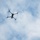 BLT 1/8 conducts organic munitions range A quadcopter drone is photographed from below against a partly cloudy blue sky. The drone is positioned in the upper-left portion of the frame, viewed from a low angle looking upward. The propellers appear to be in motion, suggesting active flight.