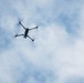 A quadcopter drone is photographed from below against a partly cloudy blue sky. The drone is positioned in the upper-left portion of the frame, viewed from a low angle looking upward. The propellers appear to be in motion, suggesting active flight.