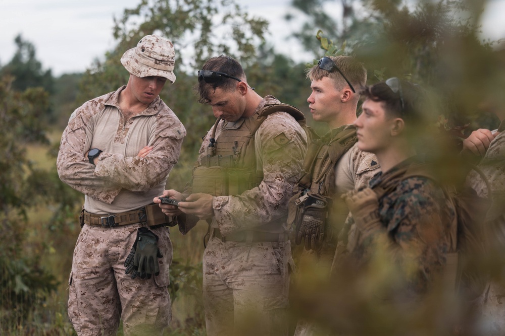 A group of military personnel in desert camouflage uniforms are gathered outdoors in a wooded or scrubland environment. One individual wearing a boonie hat appears to be instructing or observing while another examines a small handheld device, possibly a compass or GPS unit. The scene suggests a field training exercise or navigation instruction.