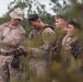 A group of military personnel in desert camouflage uniforms are gathered outdoors in a wooded or scrubland environment. One individual wearing a boonie hat appears to be instructing or observing while another examines a small handheld device, possibly a compass or GPS unit. The scene suggests a field training exercise or navigation instruction.