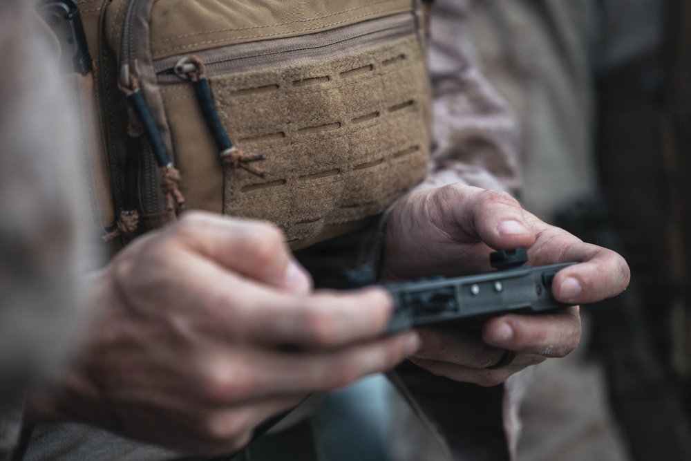 A person wearing military or tactical gear, including a tan/coyote MOLLE vest over a camouflage uniform, is handling a small black object with both hands. The object appears to be a firearm magazine or a small tactical component being inspected or loaded. The image is a close-up shot with shallow depth of field, keeping the hands and object as the focal point.