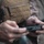 BLT 1/8 conducts organic munitions range A person wearing military or tactical gear, including a tan/coyote MOLLE vest over a camouflage uniform, is handling a small black object with both hands. The object appears to be a firearm magazine or a small tactical component being inspected or loaded. The image is a close-up shot with shallow depth of field, keeping the hands and object as the focal point.