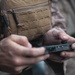 A person wearing military or tactical gear, including a tan/coyote MOLLE vest over a camouflage uniform, is handling a small black object with both hands. The object appears to be a firearm magazine or a small tactical component being inspected or loaded. The image is a close-up shot with shallow depth of field, keeping the hands and object as the focal point.