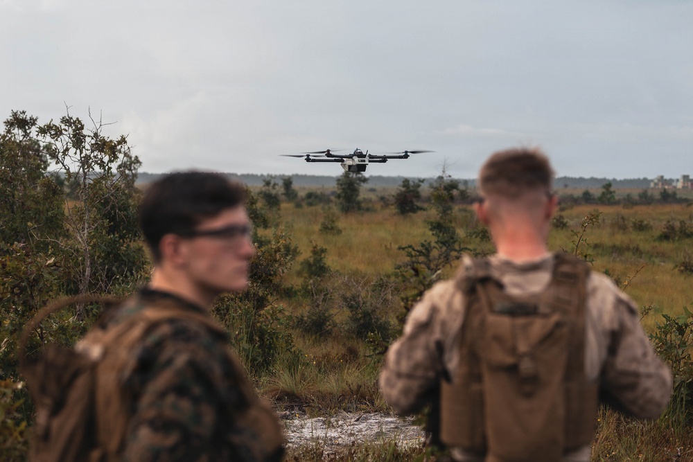 Two military personnel in camouflage uniforms observe a multi-rotor drone (hexacopter or octocopter) hovering in flight over an open scrubland terrain. The scene appears to be a field training exercise involving unmanned aerial vehicle (UAV) operations. The environment is a flat, vegetated landscape consistent with a military training range.
