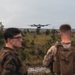 Two military personnel in camouflage uniforms observe a multi-rotor drone (hexacopter or octocopter) hovering in flight over an open scrubland terrain. The scene appears to be a field training exercise involving unmanned aerial vehicle (UAV) operations. The environment is a flat, vegetated landscape consistent with a military training range.