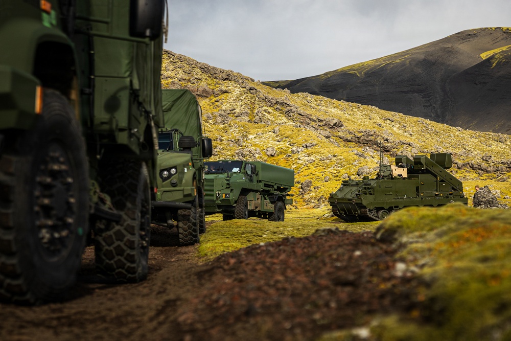 A convoy of military vehicles is positioned along a dirt track in a rugged, volcanic highland landscape with moss-covered terrain and dark basaltic hills. The vehicles are painted in olive/military green and include what appears to be wheeled transport trucks and a tracked armored vehicle with a mounted system on the right. The setting suggests a military exercise or operational deployment in a remote, likely Nordic or sub-Arctic environment.