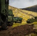 A convoy of military vehicles is positioned along a dirt track in a rugged, volcanic highland landscape with moss-covered terrain and dark basaltic hills. The vehicles are painted in olive/military green and include what appears to be wheeled transport trucks and a tracked armored vehicle with a mounted system on the right. The setting suggests a military exercise or operational deployment in a remote, likely Nordic or sub-Arctic environment.