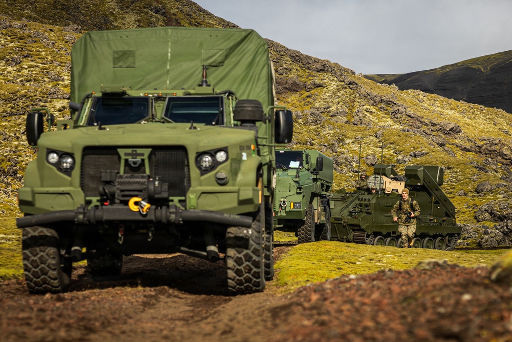 A military convoy of green tactical vehicles is positioned in a rugged, moss-covered highland terrain, likely Iceland based on the distinctive volcanic landscape. The scene includes at least two wheeled vehicles and one tracked vehicle, with uniformed personnel visible near the equipment. This appears to be a military exercise or operational deployment in a remote environment.