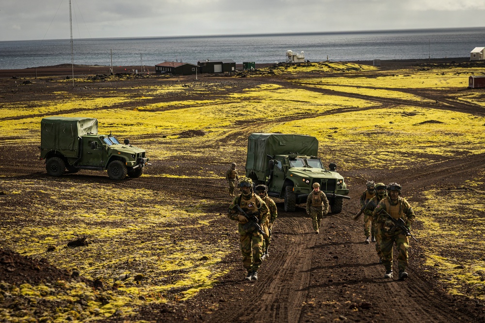 A group of armed military personnel in camouflage uniforms advance on foot along a dirt track, accompanied by two green military tactical vehicles (appearing to be Oshkosh JLTV-type trucks) with canvas-covered cargo beds. The terrain is characterized by dark volcanic soil with patches of bright yellow-green moss or lichen, suggesting a Nordic or sub-Arctic environment, likely Iceland. Military infrastructure including antenna towers, radar domes, and small buildings is visible in the background near a coastline.