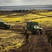 A group of armed military personnel in camouflage uniforms advance on foot along a dirt track, accompanied by two green military tactical vehicles (appearing to be Oshkosh JLTV-type trucks) with canvas-covered cargo beds. The terrain is characterized by dark volcanic soil with patches of bright yellow-green moss or lichen, suggesting a Nordic or sub-Arctic environment, likely Iceland. Military infrastructure including antenna towers, radar domes, and small buildings is visible in the background near a coastline.