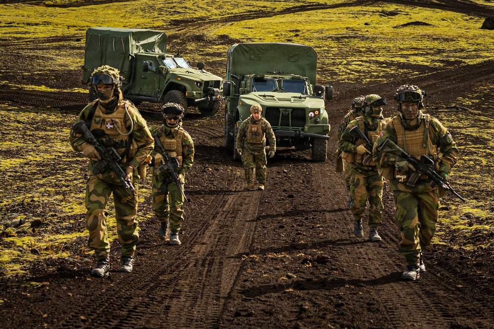 A group of five military personnel in camouflage uniforms are walking forward along a dirt track in a volcanic or moorland terrain with bright green moss-covered ground. Two military tactical vehicles with green canvas-covered cargo beds are parked or staged behind them. The soldiers are armed and equipped with tactical gear, suggesting a training exercise or operational patrol.