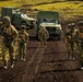 A group of five military personnel in camouflage uniforms are walking forward along a dirt track in a volcanic or moorland terrain with bright green moss-covered ground. Two military tactical vehicles with green canvas-covered cargo beds are parked or staged behind them. The soldiers are armed and equipped with tactical gear, suggesting a training exercise or operational patrol.