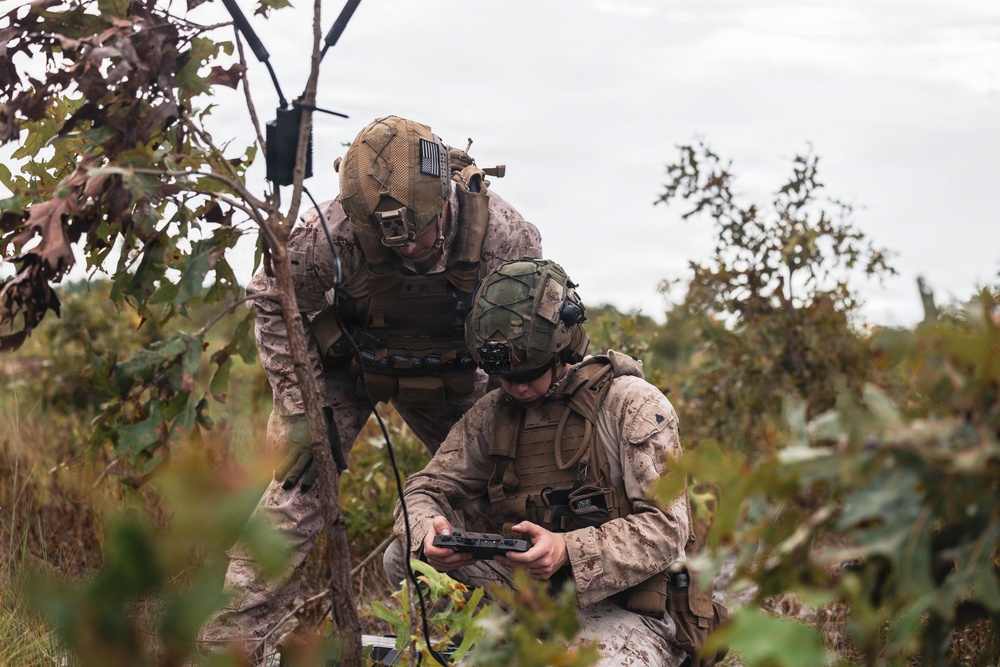 Two military personnel in desert MARPAT camouflage uniforms are crouched in a vegetated field, collaborating over a handheld electronic device, likely a tablet or drone controller. Both wear tactical helmets with mounted accessories and body armor with MOLLE gear. The setting appears to be an outdoor training or operational environment with sparse shrubs and an overcast sky.