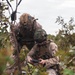 Two military personnel in desert MARPAT camouflage uniforms are crouched in a vegetated field, collaborating over a handheld electronic device, likely a tablet or drone controller. Both wear tactical helmets with mounted accessories and body armor with MOLLE gear. The setting appears to be an outdoor training or operational environment with sparse shrubs and an overcast sky.