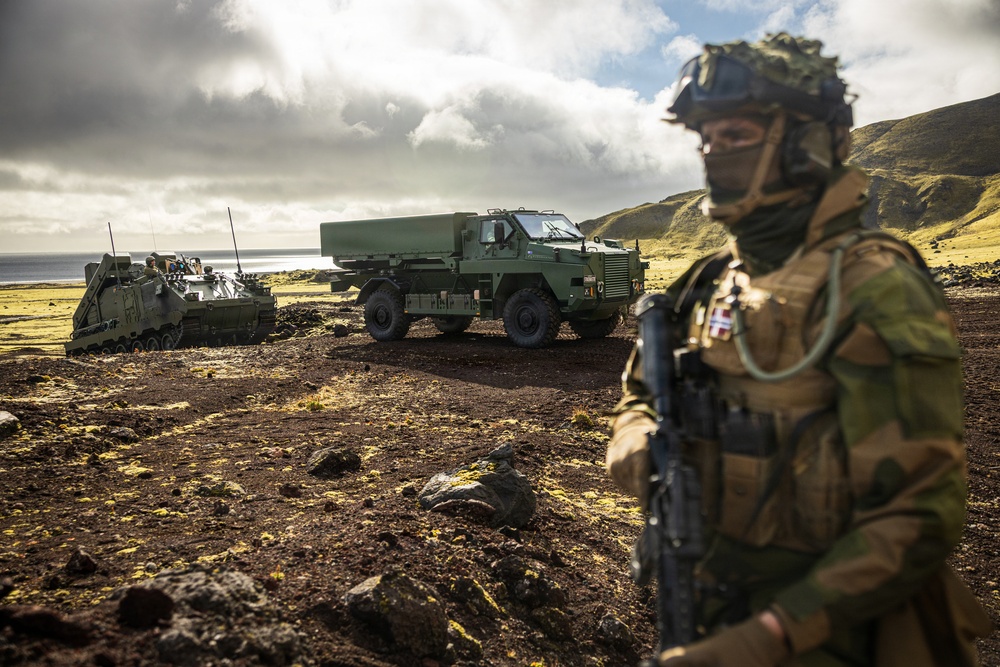 A fully equipped soldier in camouflage gear stands in the foreground, wearing a flag patch consistent with the Danish or Norwegian flag on his shoulder. Behind him are two military vehicles — what appears to be an armored personnel carrier and a large green tactical truck — positioned on rocky, volcanic-looking terrain near a coastline. The setting and landscape suggest a Nordic or North Atlantic environment, possibly Iceland.