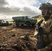 A fully equipped soldier in camouflage gear stands in the foreground, wearing a flag patch consistent with the Danish or Norwegian flag on his shoulder. Behind him are two military vehicles — what appears to be an armored personnel carrier and a large green tactical truck — positioned on rocky, volcanic-looking terrain near a coastline. The setting and landscape suggest a Nordic or North Atlantic environment, possibly Iceland.
