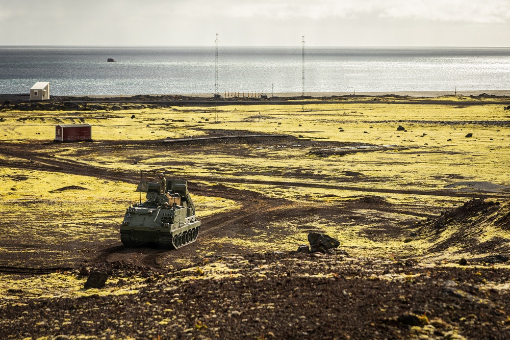 A military tracked armored personnel carrier (likely an M113 or variant) is traversing a rugged, volcanic coastal terrain with yellow-green moss-covered ground. The landscape appears to be a remote, sparsely populated coastal area with antenna towers and small structures visible in the background near the shoreline. The scene suggests a military training exercise or patrol operation in a Nordic or sub-Arctic environment.