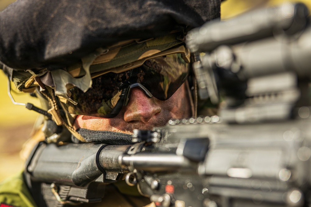 A close-up photograph of a military or tactical operator in a prone or low shooting position, aiming down the sights of a firearm. The subject is wearing camouflage gear, a helmet with netting and foliage, and ballistic/tactical eyewear. The image is tightly framed on the face and weapon, with a shallow depth of field creating bokeh in the background.
