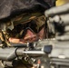 A close-up photograph of a military or tactical operator in a prone or low shooting position, aiming down the sights of a firearm. The subject is wearing camouflage gear, a helmet with netting and foliage, and ballistic/tactical eyewear. The image is tightly framed on the face and weapon, with a shallow depth of field creating bokeh in the background.