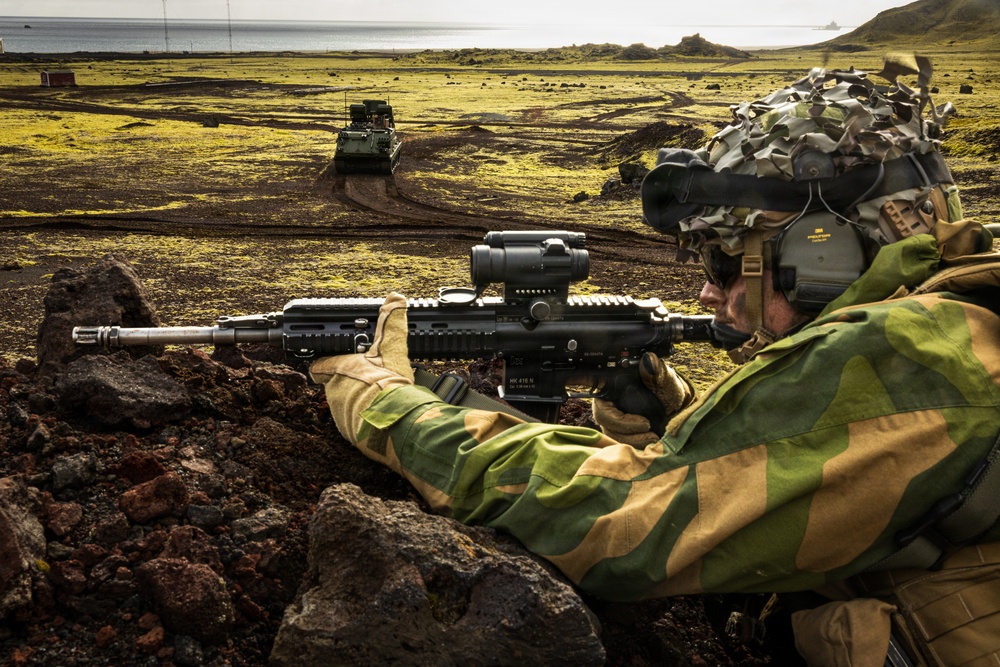 A soldier in camouflage gear is positioned behind volcanic rocks in a prone/crouched firing stance, aiming a rifle equipped with an optical scope. The terrain appears to be a volcanic coastal landscape, likely Iceland, with moss-covered lava fields visible in the background. An armored military vehicle is visible in the mid-ground, suggesting a military exercise or operation.