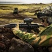 A soldier in camouflage gear is positioned behind volcanic rocks in a prone/crouched firing stance, aiming a rifle equipped with an optical scope. The terrain appears to be a volcanic coastal landscape, likely Iceland, with moss-covered lava fields visible in the background. An armored military vehicle is visible in the mid-ground, suggesting a military exercise or operation.