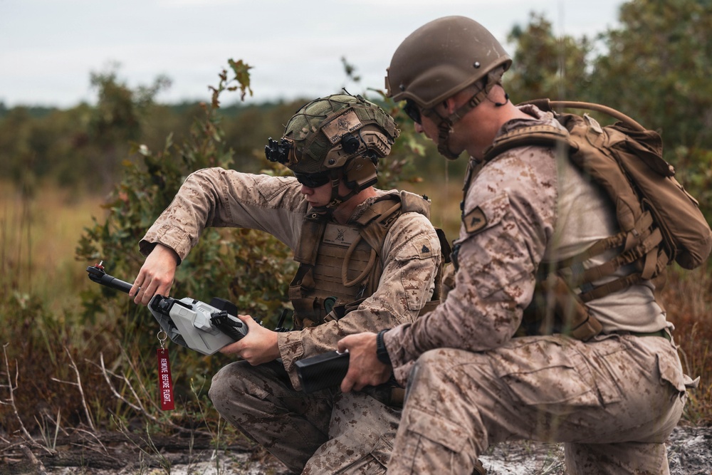 Two military personnel in desert camouflage uniforms are crouched in an outdoor field environment, examining what appears to be a drone controller or remote control device. Both individuals are wearing tactical gear including helmets, body armor, and equipment vests. The setting appears to be a training exercise or field operation in a scrubland environment.